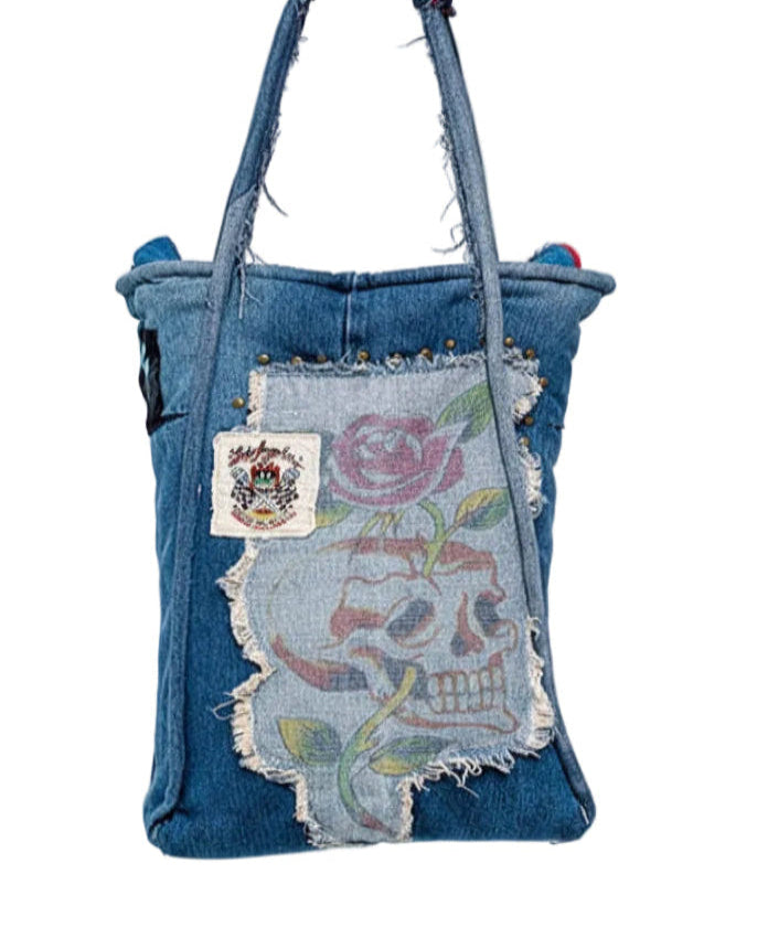 A blue denim tote bag featuring a distressed front patch with a floral skull design and a small embroidered brand patch.