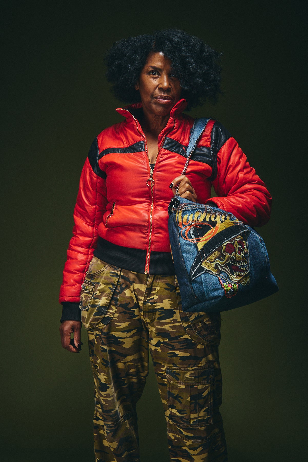 Person wearing a red puffer jacket and camouflage pants holding a colorful bag against a dark background